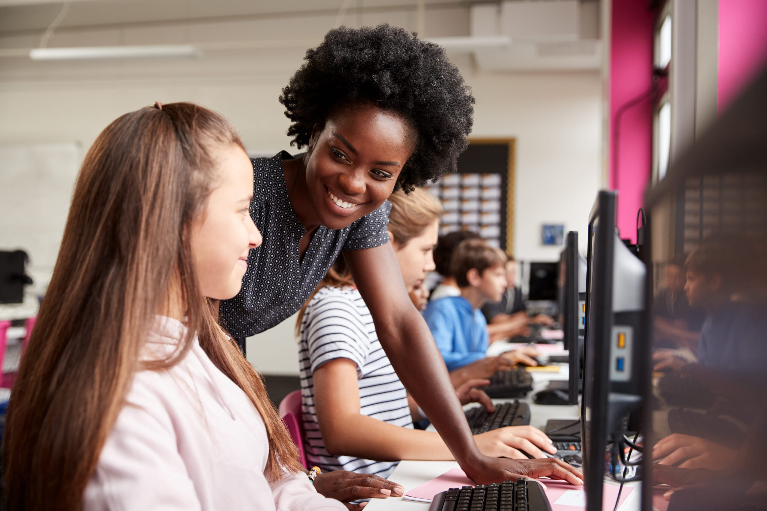 Instructor assisting a student at a desktop computer while other students work in a computer lab.