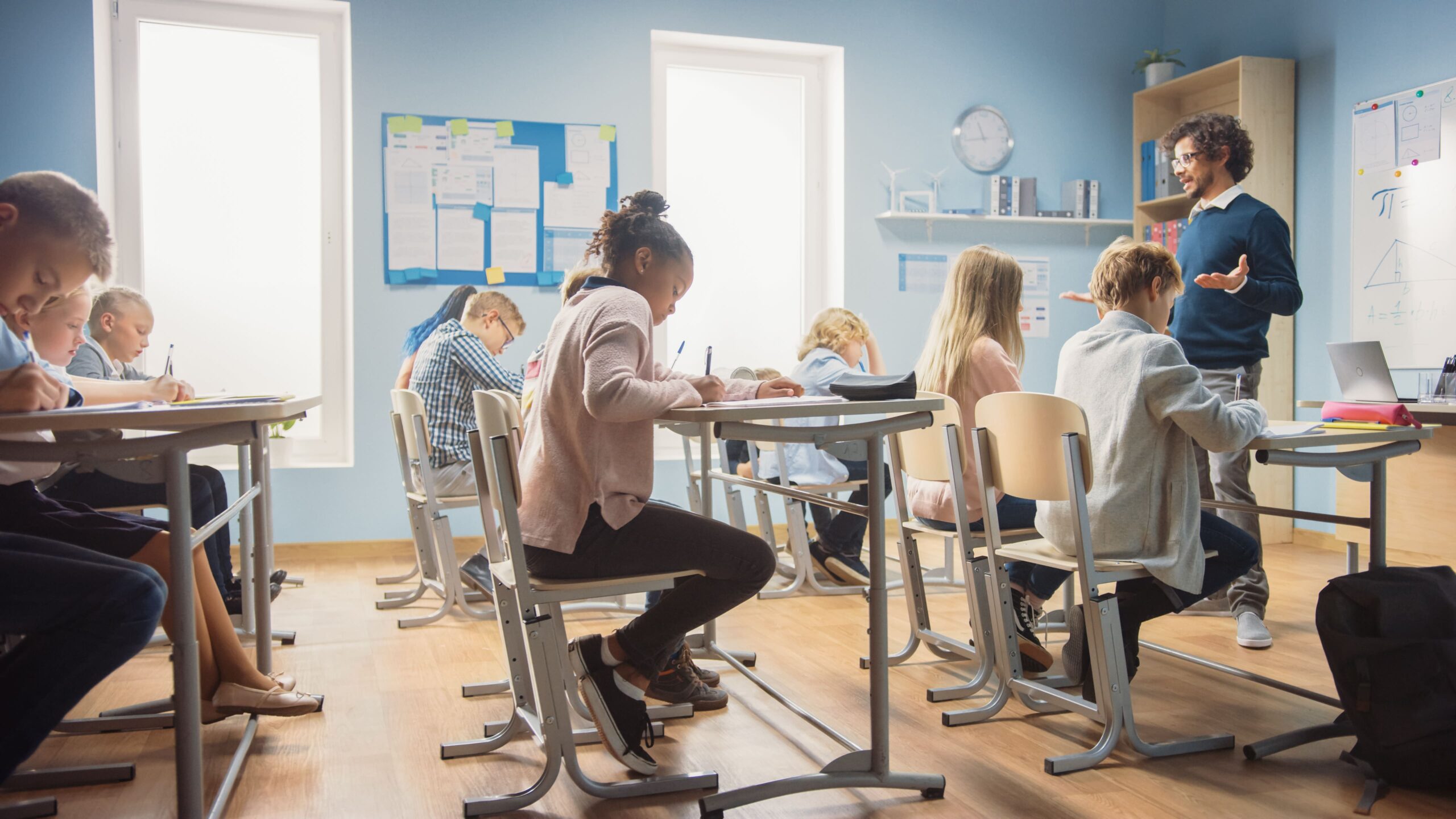 Students seated at desks writing while an instructor stands at the front near a whiteboard.