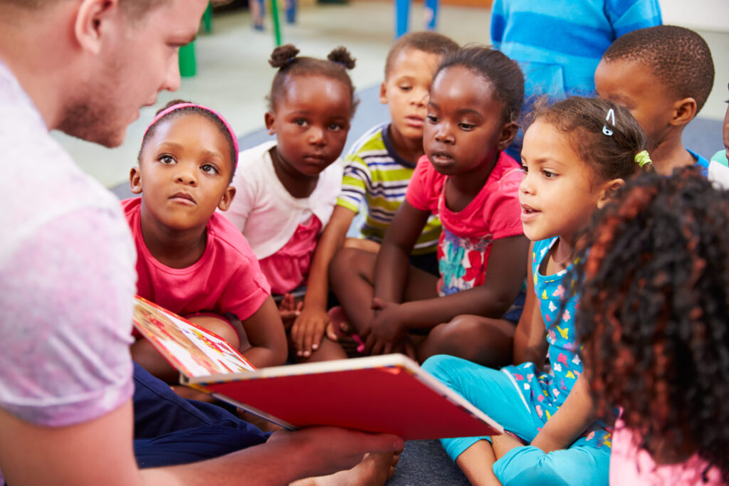 Adult reading a book to a group of children seated closely together on the floor.