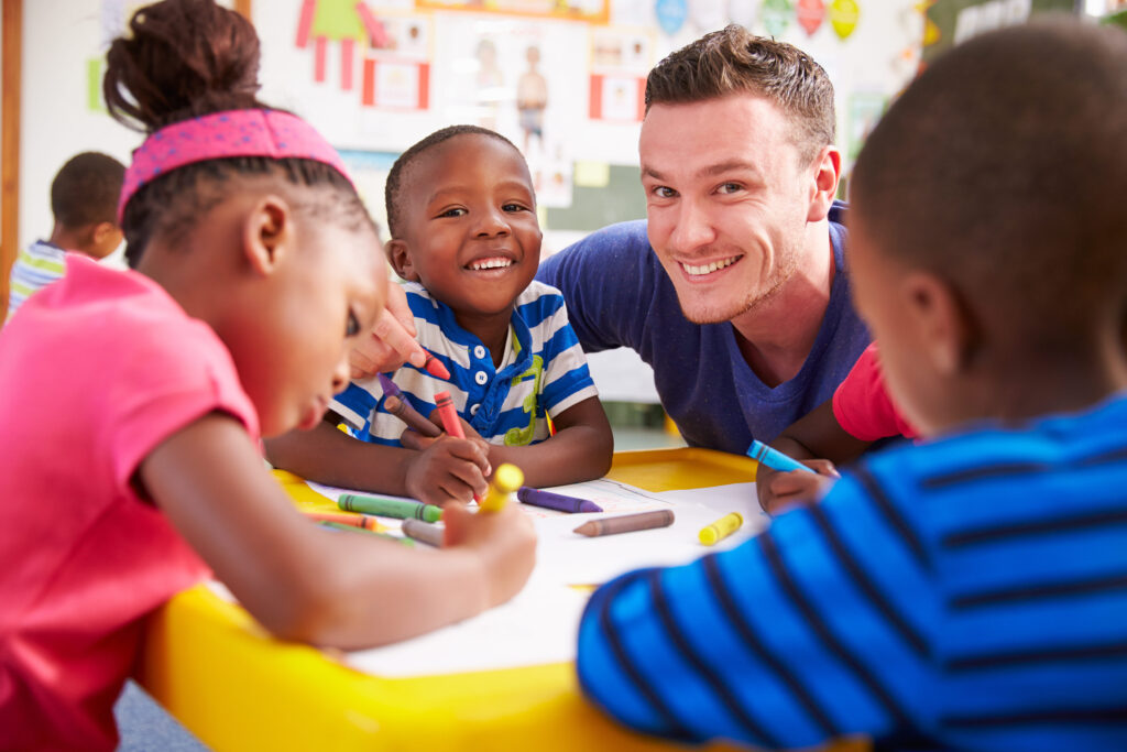 Children coloring with crayons at a table while an adult sits with them.
