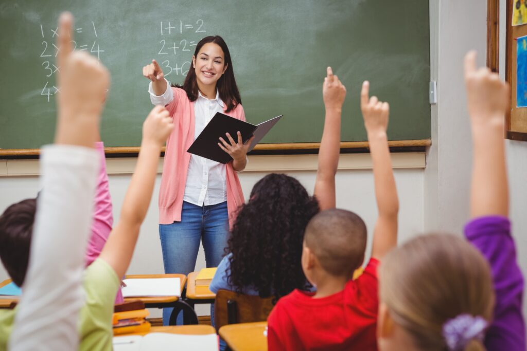 Teacher pointing at a student while several students raise their hands in a classroom.